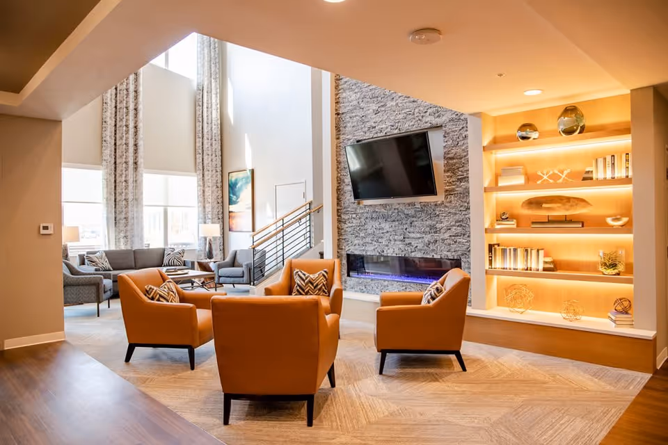 A modern living room area in The Lodge at Greeley featuring four orange armchairs arranged around a central point, a stone wall with a mounted flat-screen TV above a sleek electric fireplace, and built-in illuminated shelves displaying decorative items and books. Large windows with patterned curtains allow natural light to fill the space, and additional seating with gray sofas and chairs is visible in the background.