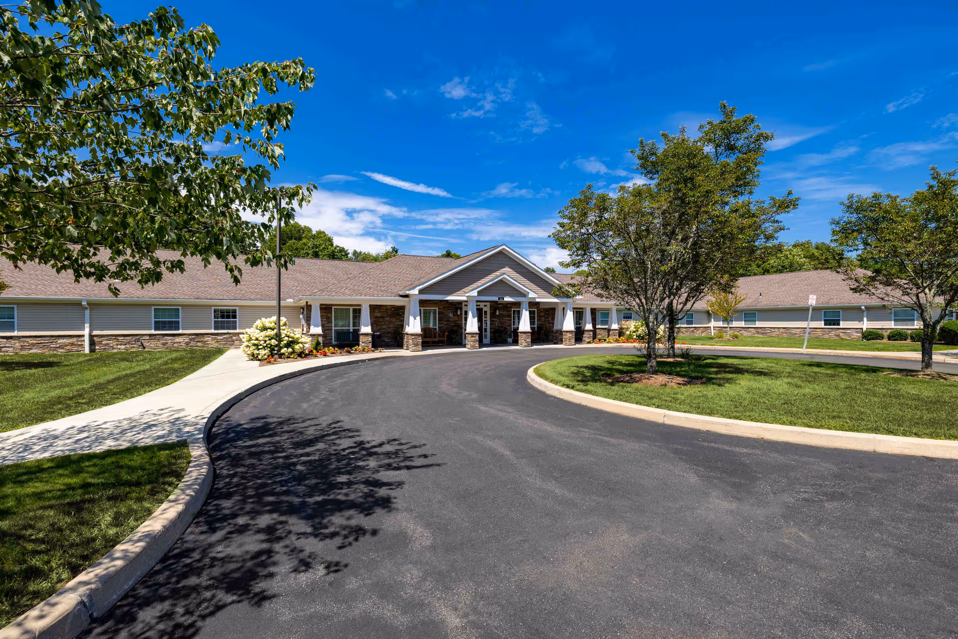 Sunlit senior living facility with circular driveway and landscaped entrance under blue sky.