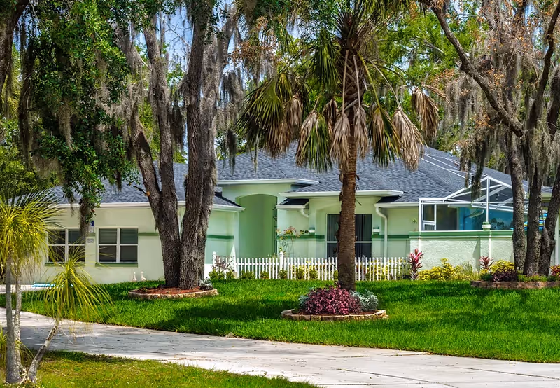 Exterior view of a green assisted living facility with palm trees and lawn.