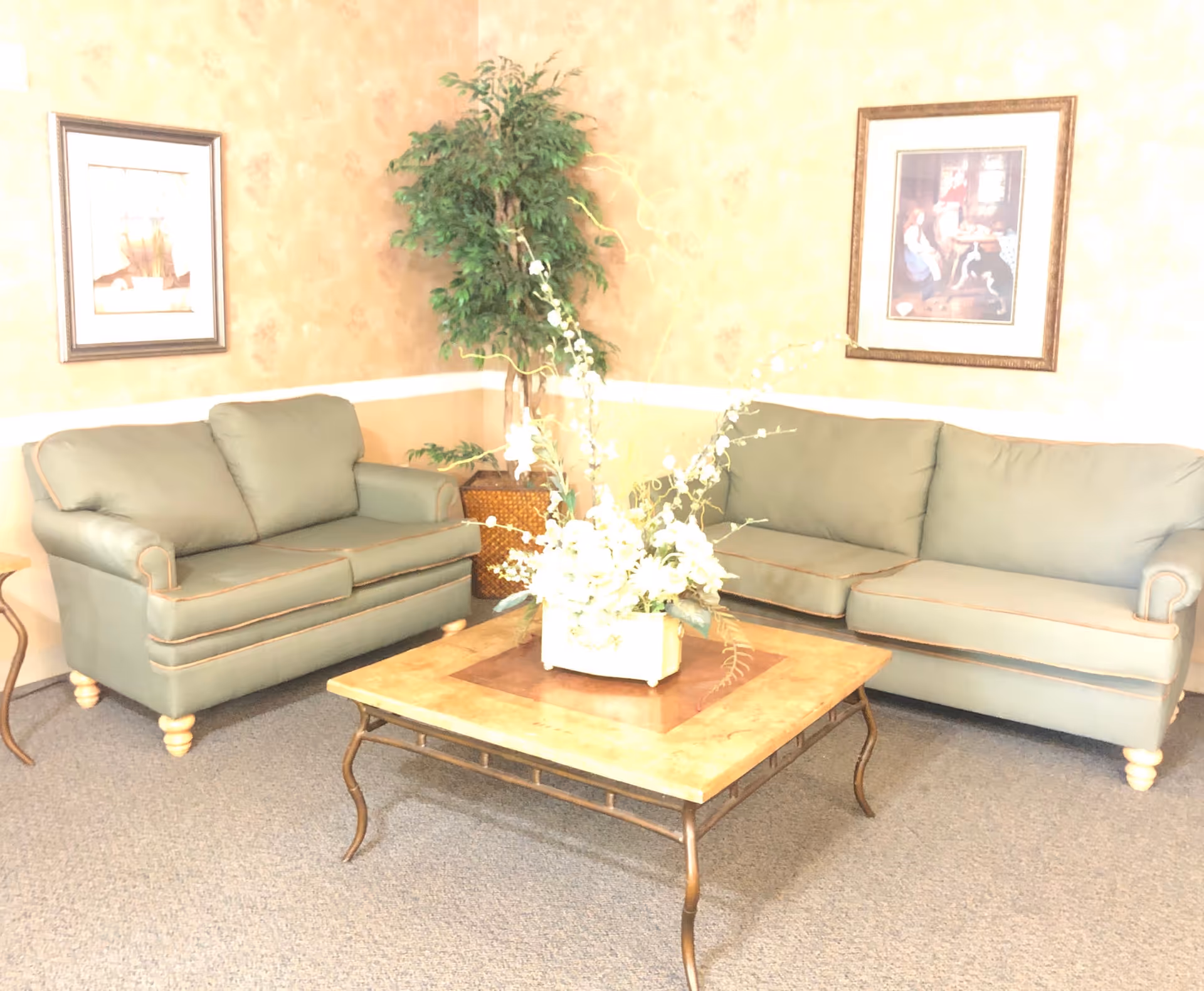 A seating area with two green sofas, a wooden coffee table with a floral centerpiece, framed pictures, and a potted plant.