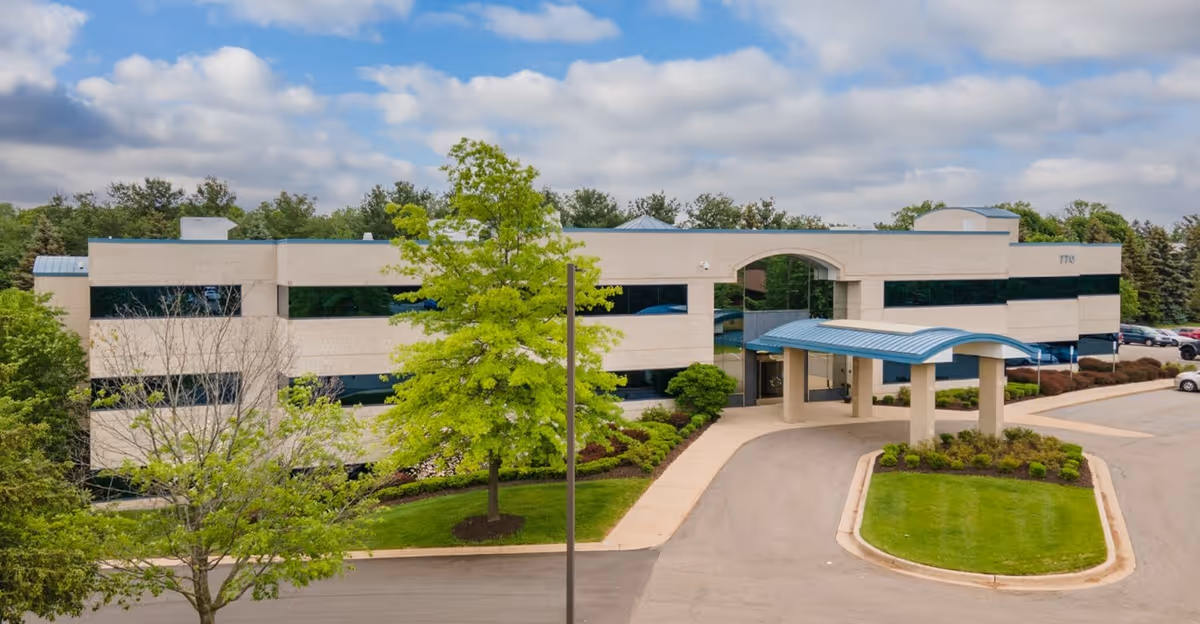 A modern two-story senior living facility exterior with curved entrance canopy and greenery.