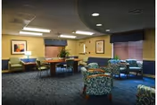 A cozy senior facility common room with blue chairs, tables, lamps, and artwork.