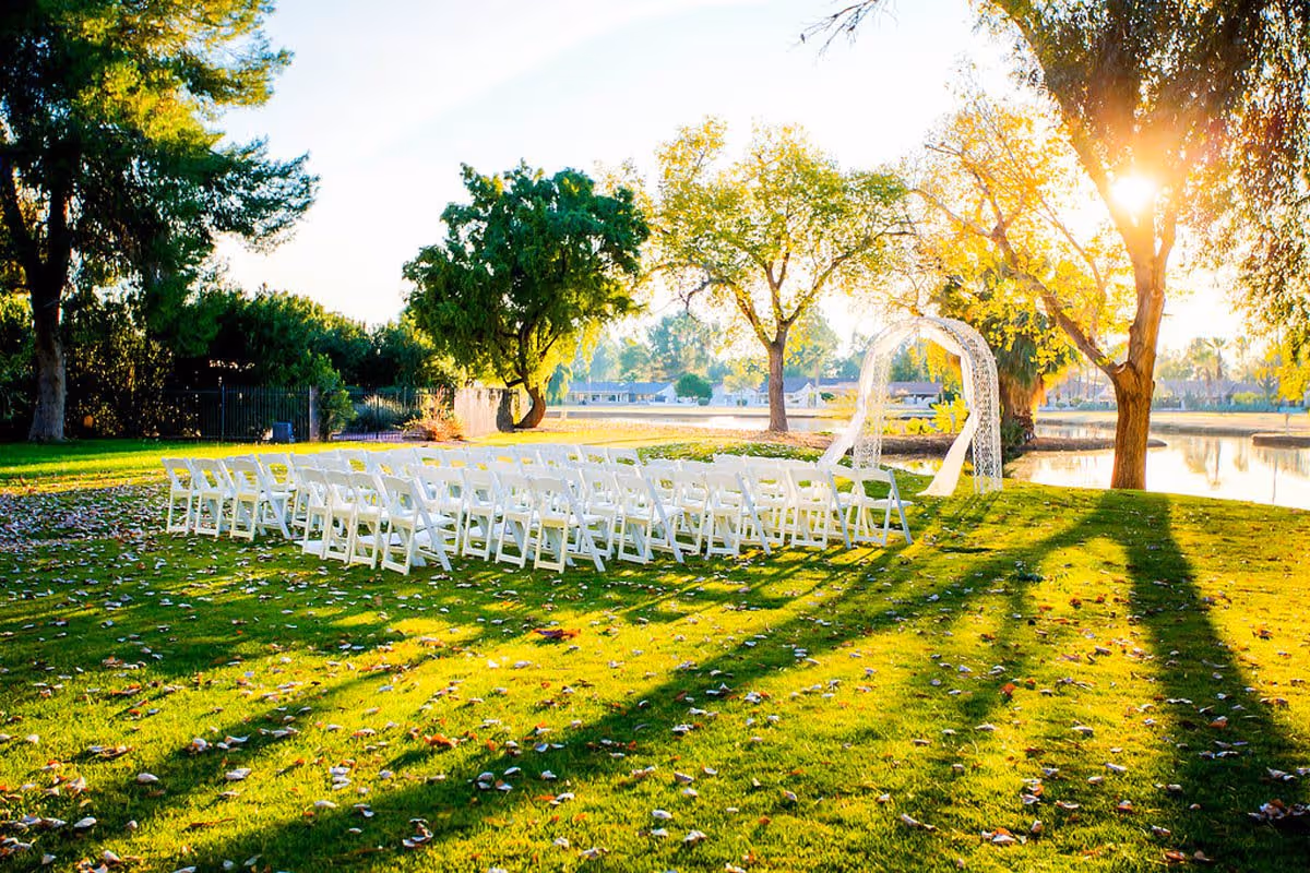 Rows of white folding chairs and a decorative arch set up on a sunlit grassy lawn beside a lake and trees.
