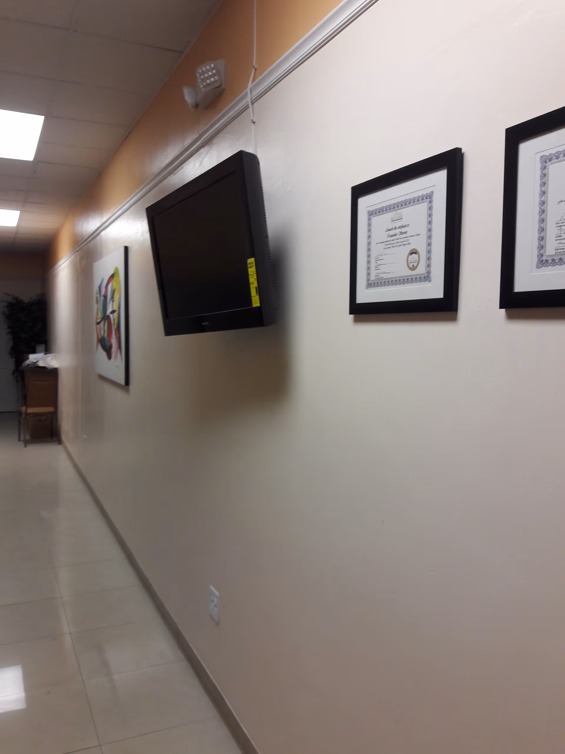 A long beige hallway with a wall-mounted TV and framed certificates on the wall.