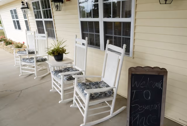 Front porch of a yellow-sided building with three white rocking chairs, a small table with a potted plant, and a chalkboard welcome sign.