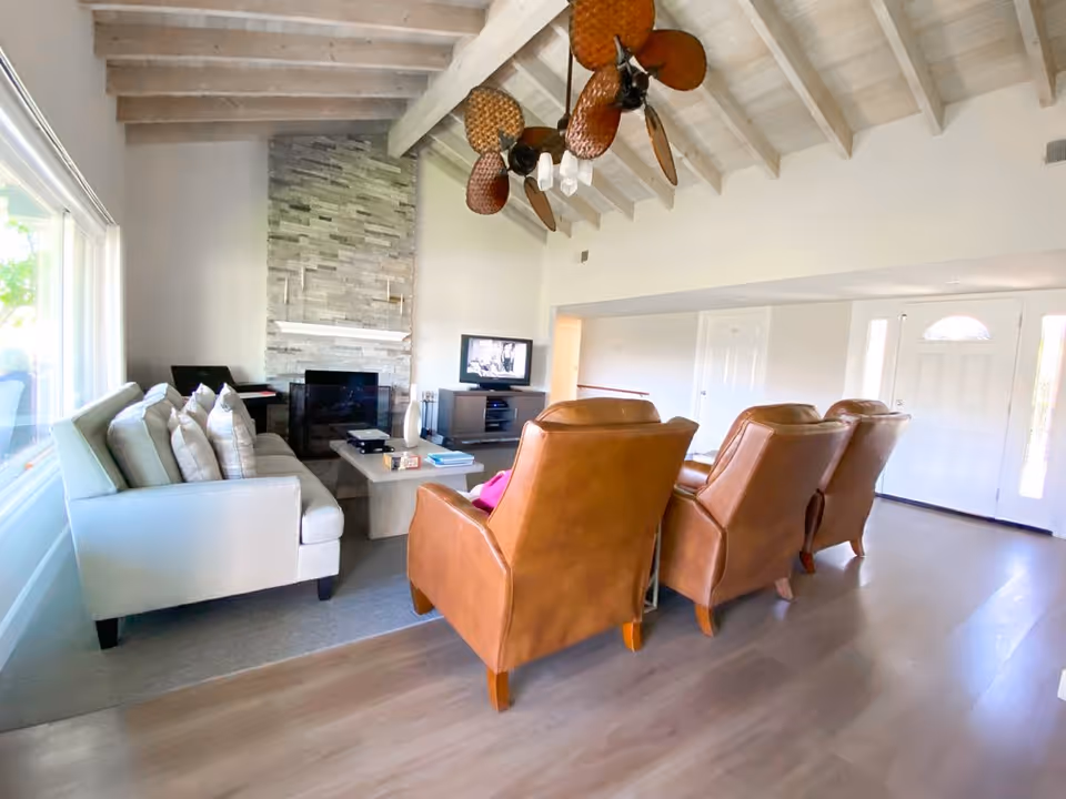 Bright living room with leather chairs, a sofa, and a TV beside a stone fireplace.