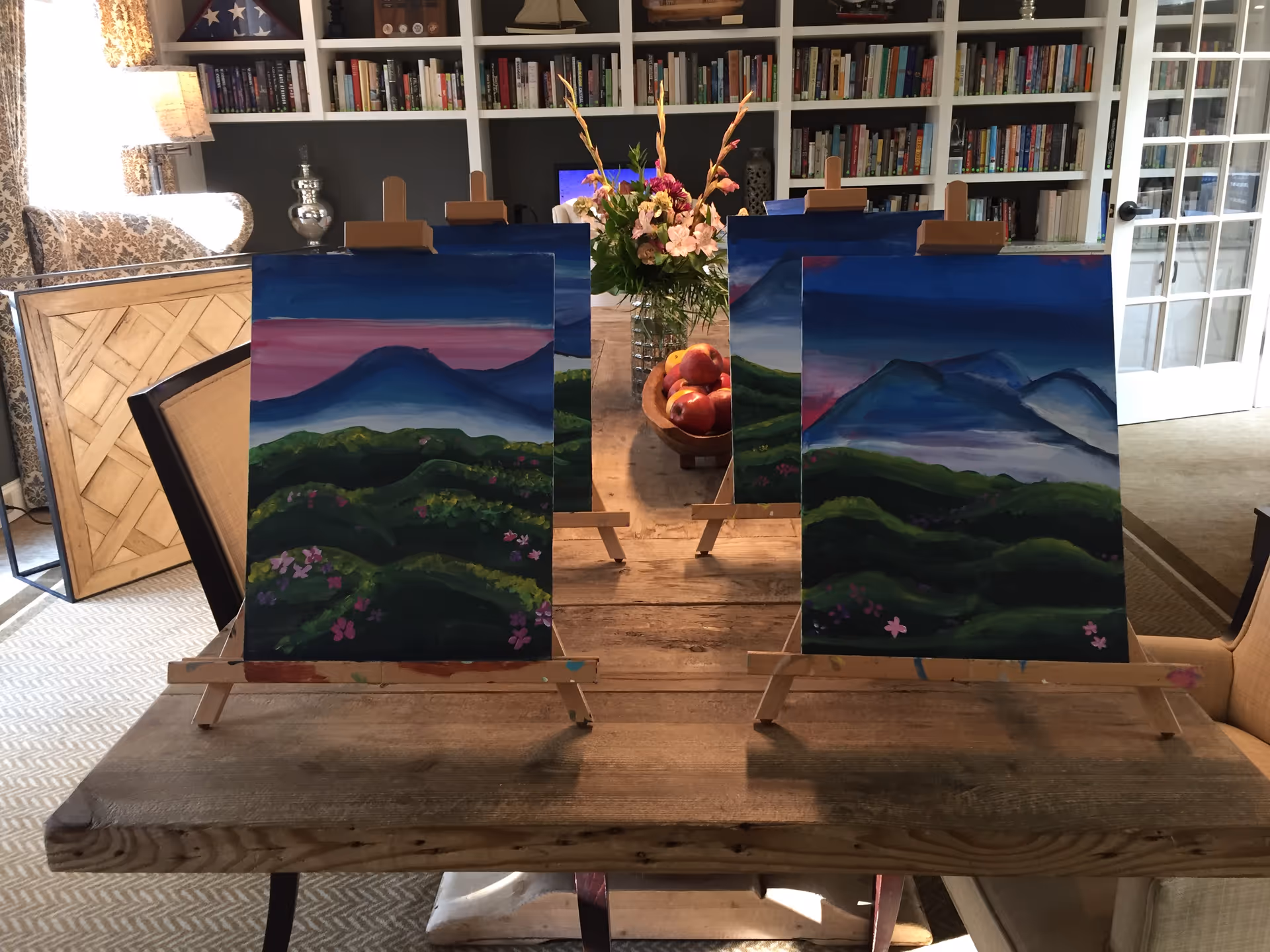 Two landscape paintings on easels sit on a rustic table in a book-filled living room.