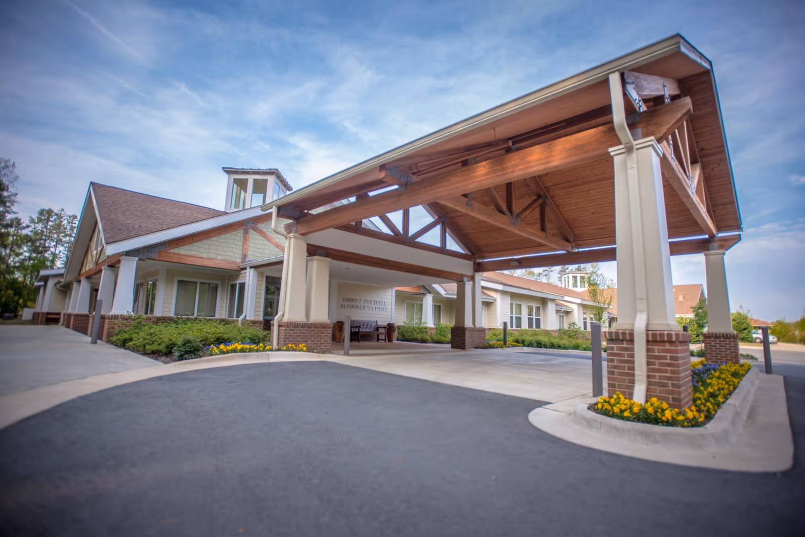 Entrance of a single-story senior living facility with a large wooden porte-cochere, brick columns, and landscaped flowerbeds.