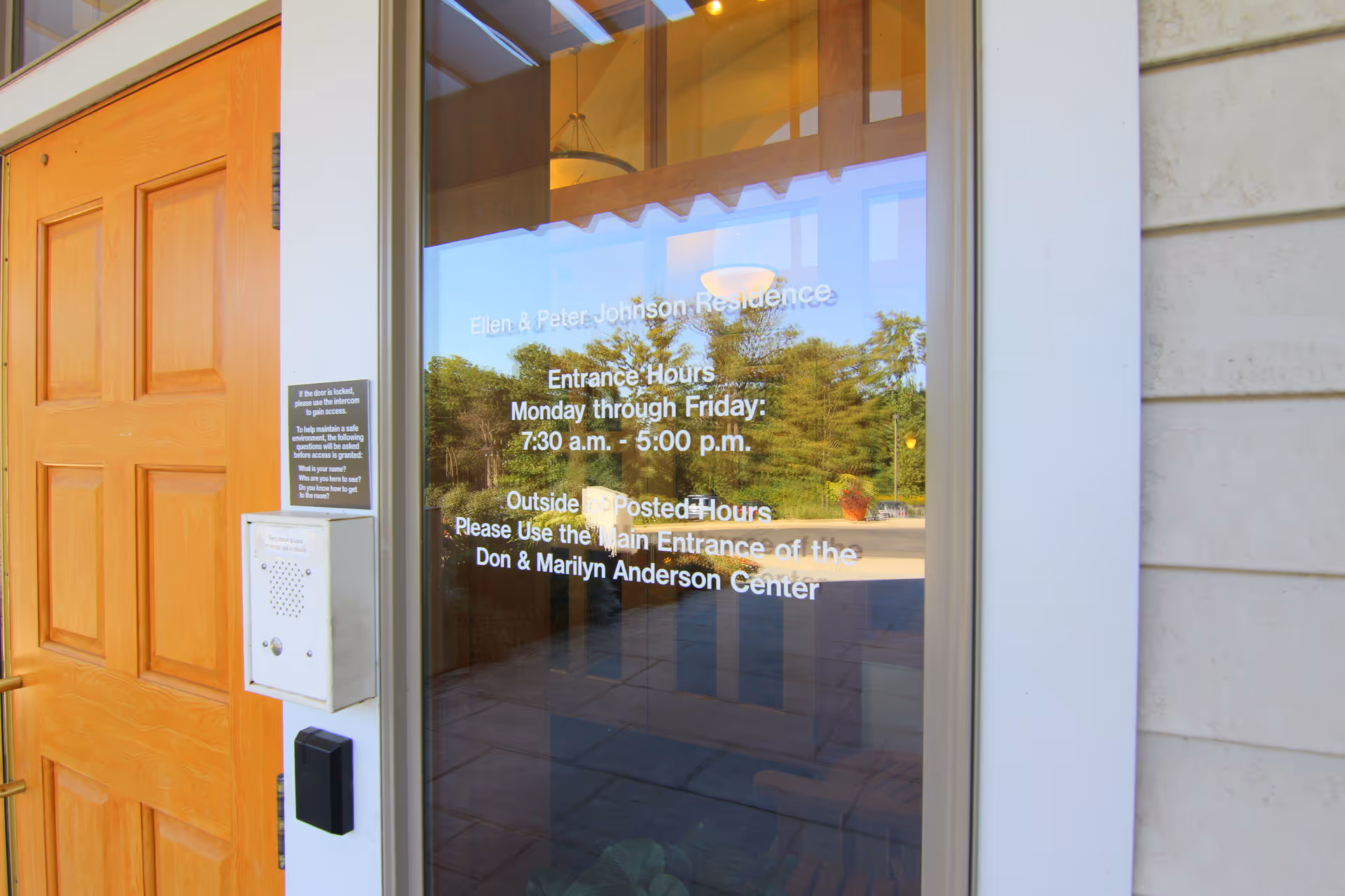 Glass entrance with hours text reflecting trees beside a wooden door.