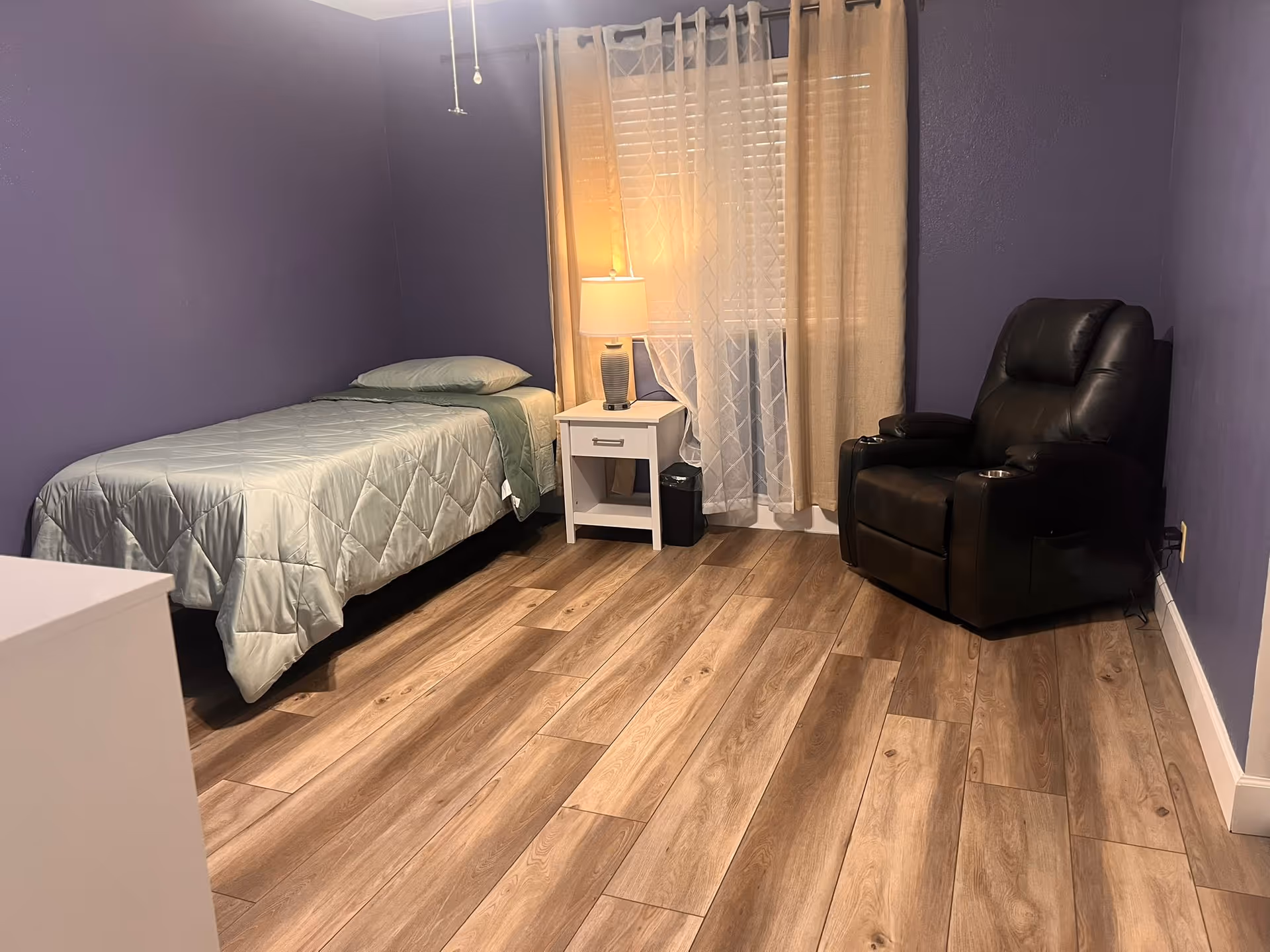 Purple-walled bedroom with a single bed, nightstand, lamp, and a black recliner.