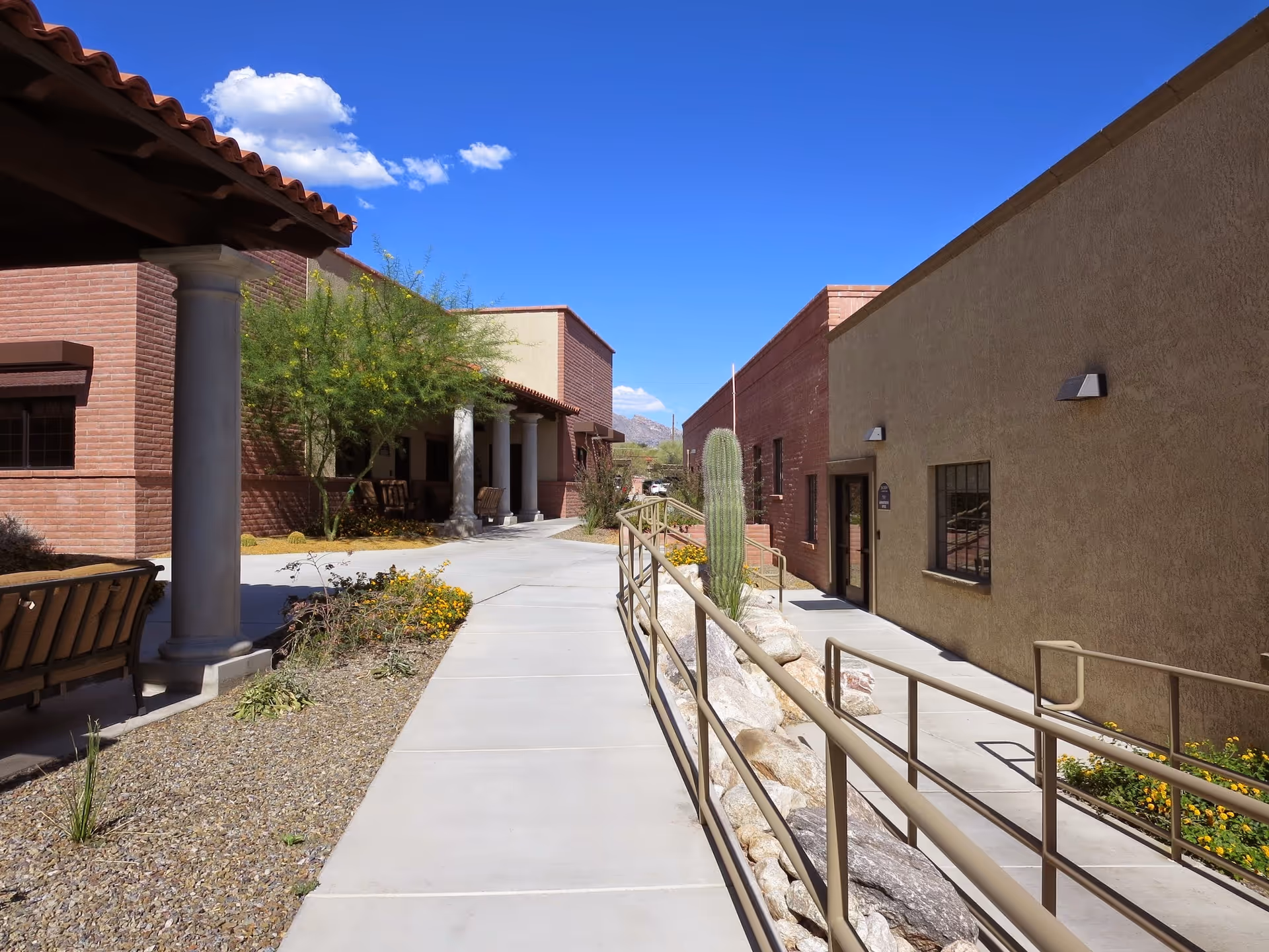 Sunny desert courtyard between beige buildings with walkway, railing, cacti, and benches.