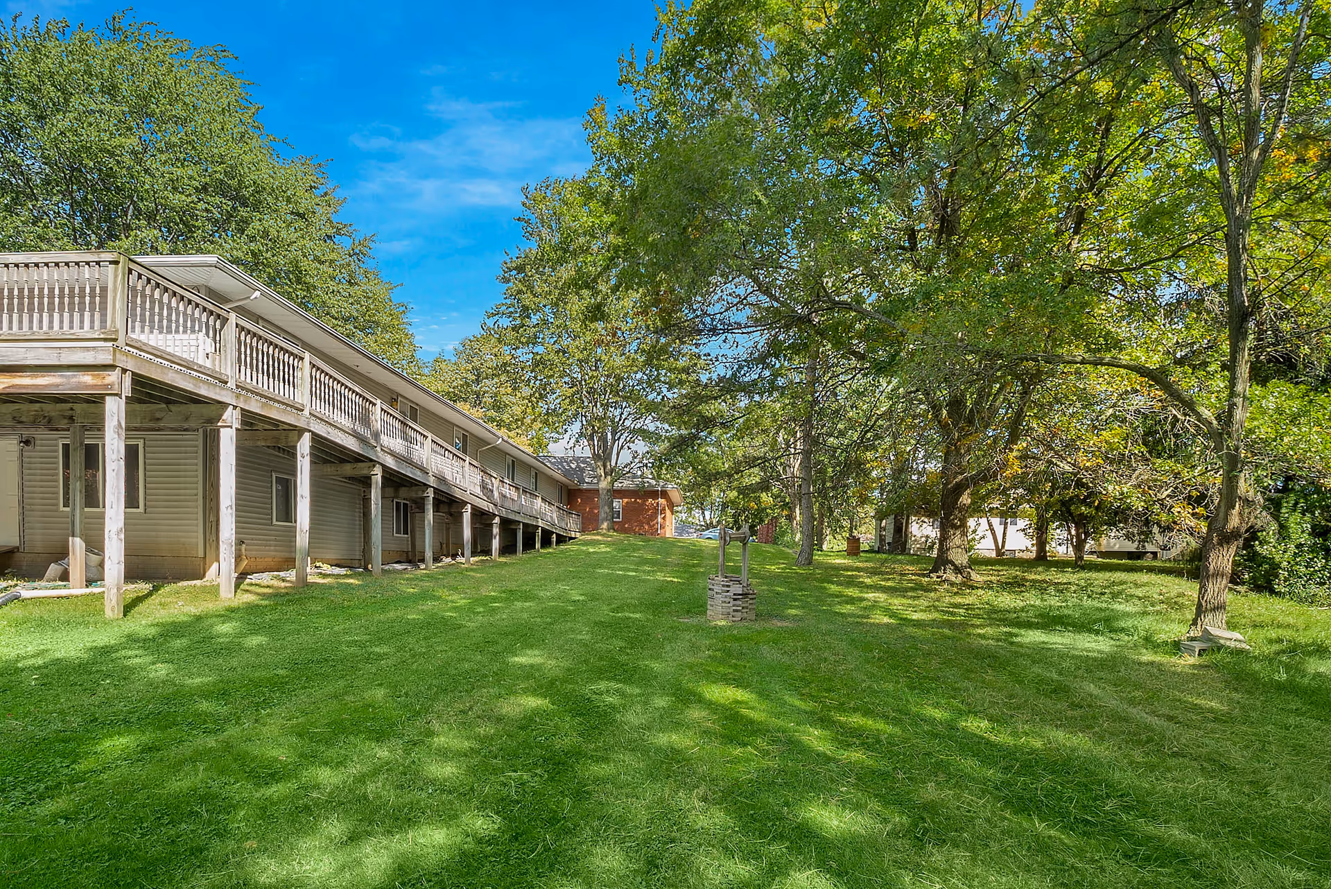 Outdoor view of a senior living facility with a grassy lawn, several trees, and a long building with a wooden balcony on the left side under a clear blue sky.