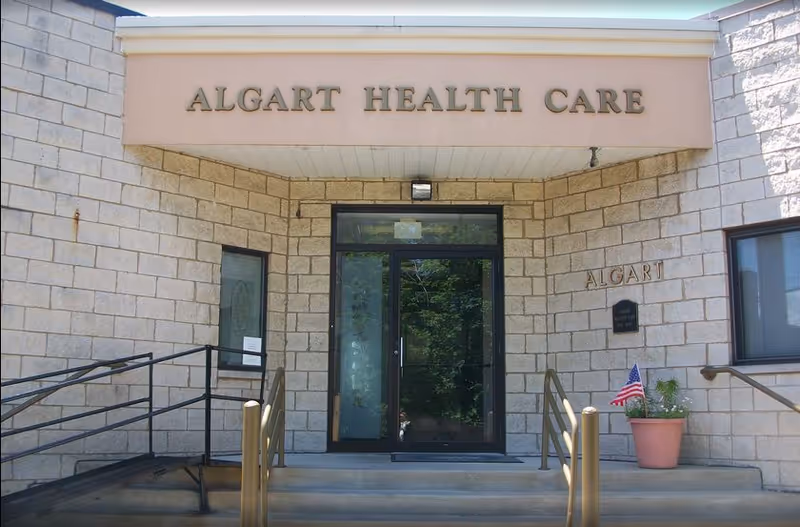 Main entrance of Algart Health Care with glass double doors set in a stone facade, steps and handrails, and a potted American flag.