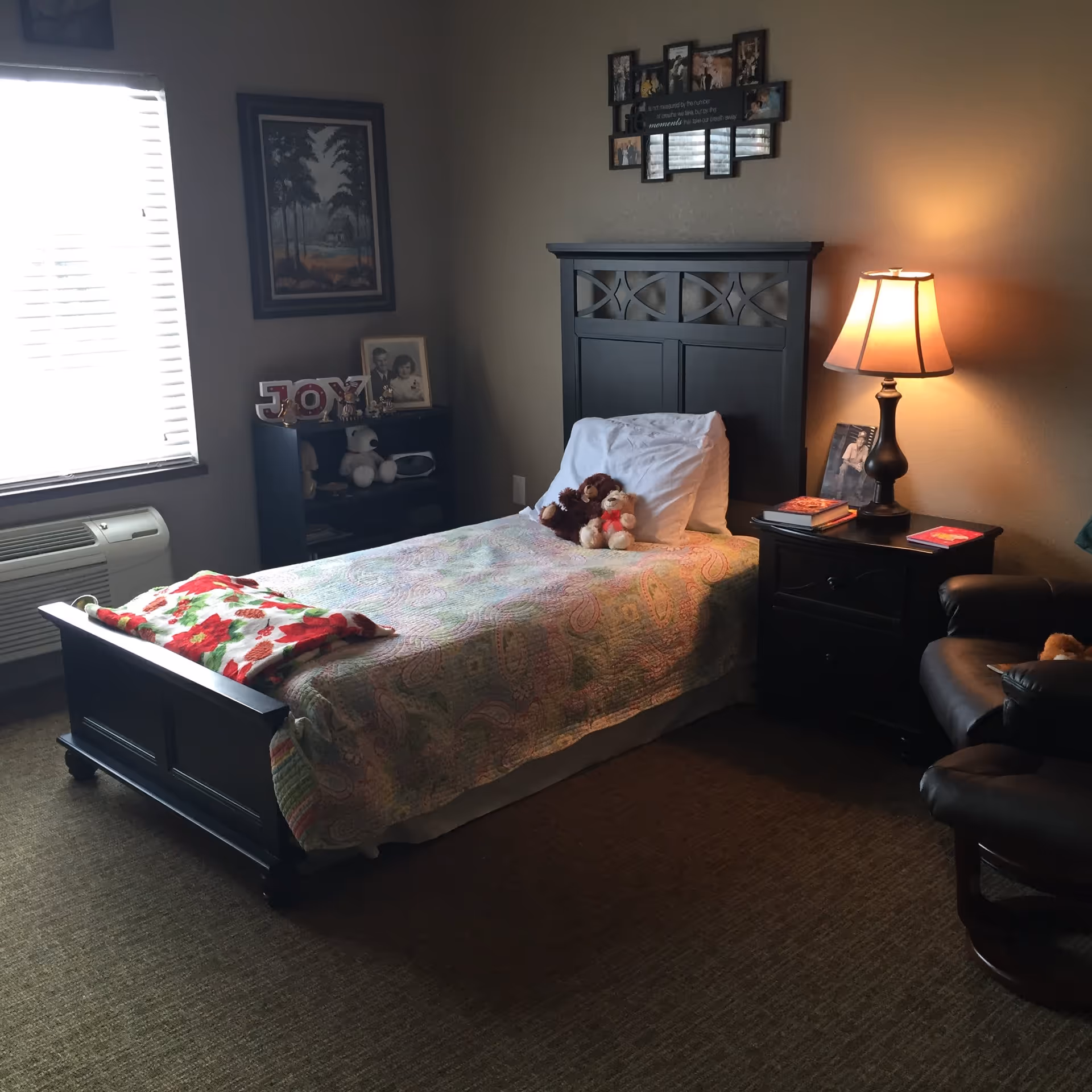 A cozy bedroom with a single bed covered in a colorful quilt and two stuffed animals on the pillow. Next to the bed is a dark wooden nightstand with a lamp, books, and a framed photo. There is a dark wooden bookshelf with decorative items and a framed photo near a window with blinds. A comfortable leather chair is partially visible on the right side. The walls are decorated with framed pictures and a photo collage.