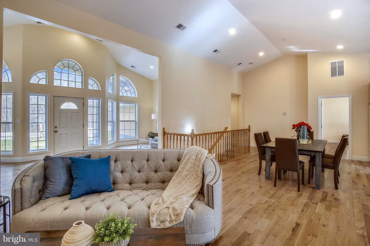 A spacious living and dining area with light wood flooring and beige walls. The living room features a tufted beige sofa with two pillows and a throw blanket, positioned near large windows with decorative arches that let in natural light. The dining area has a dark wooden table with six matching chairs and a centerpiece with red flowers. Recessed ceiling lights illuminate the space.