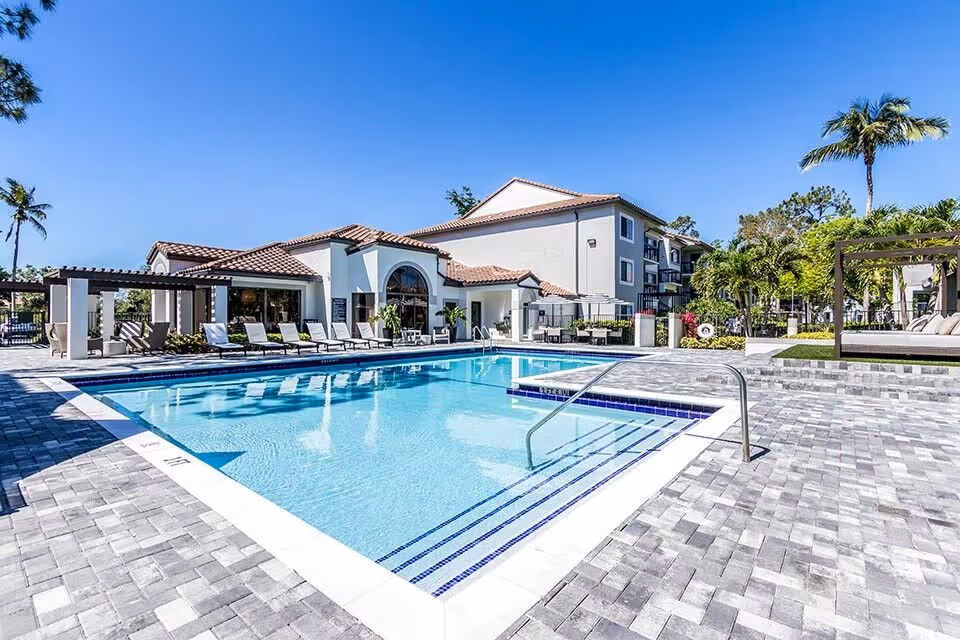 Outdoor swimming pool area at a senior living facility with lounge chairs lined up along the poolside, a pergola with seating, palm trees, and a multi-story building in the background under a clear blue sky.