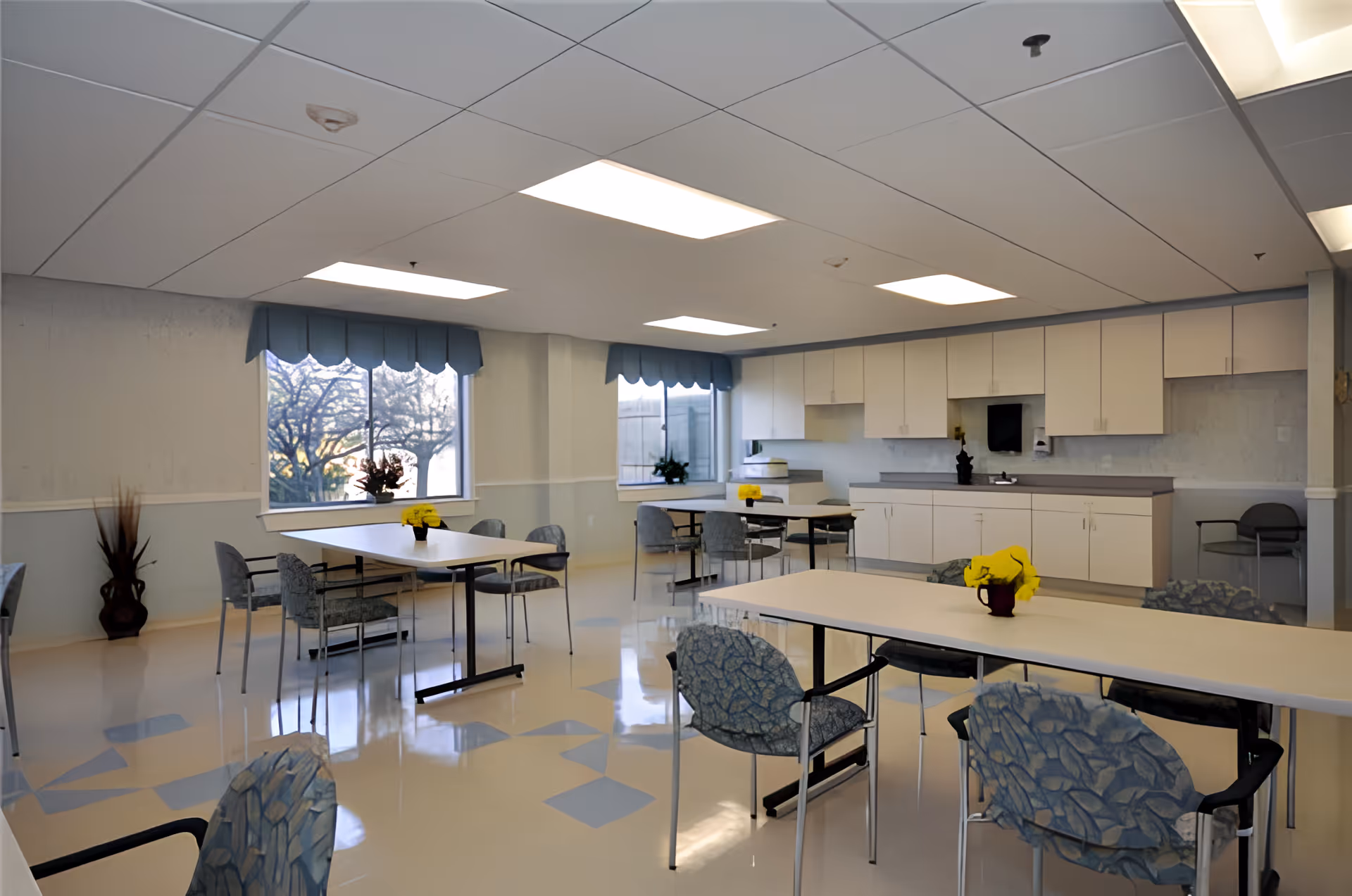 Bright communal dining/activity room with tables and patterned chairs and a kitchenette along the back wall.
