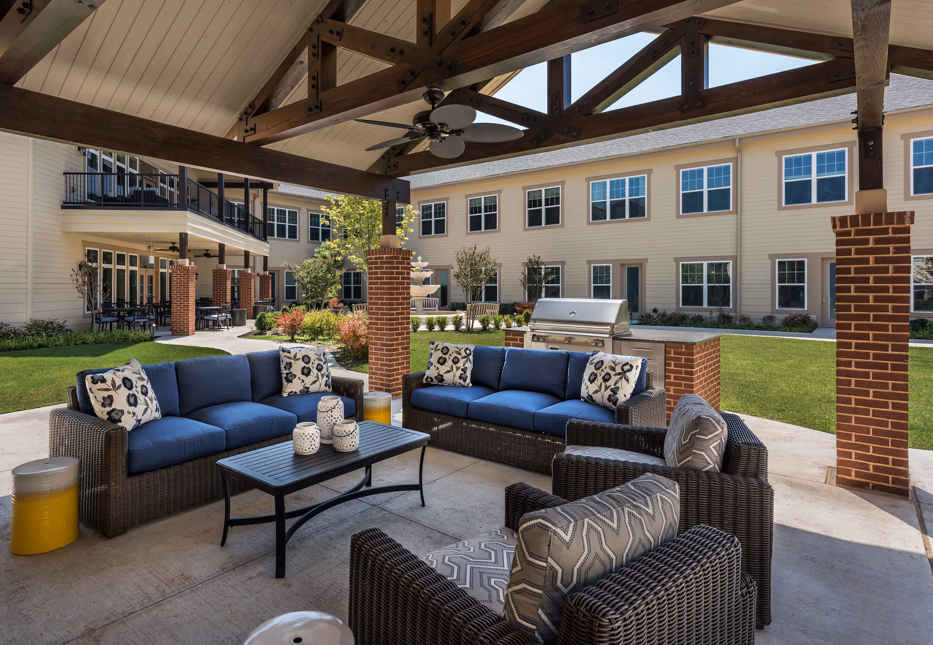 Outdoor covered seating area with wicker furniture including two blue cushioned sofas and two armchairs with patterned cushions, a black coffee table with decorative items, and a built-in grill. The area is shaded by a wooden pergola with ceiling fans. In the background, there is a two-story building with multiple windows and doors, surrounded by green grass and landscaping.