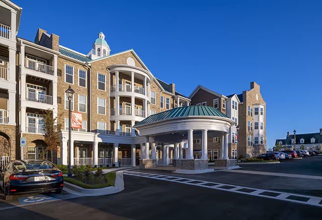 Multistory senior living building with a circular covered porte-cochère entrance, balconies, and parked cars under a clear blue sky.