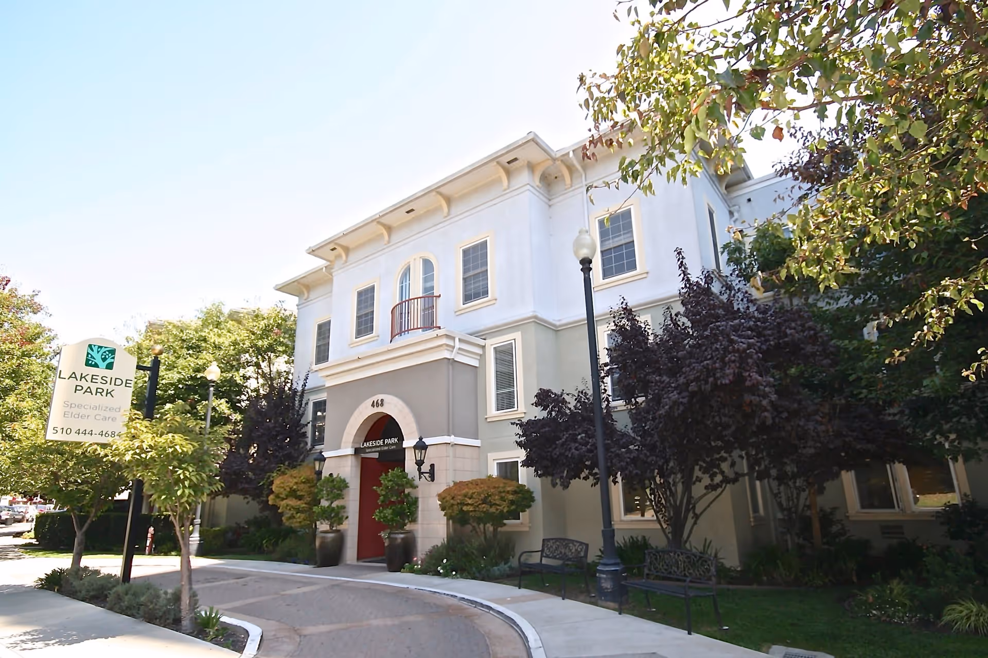 Exterior view of Lakeside Park, a specialized elder care facility, showing a three-story building with a red entrance door, surrounded by trees and greenery. There is a sign near the sidewalk with the facility name and phone number.