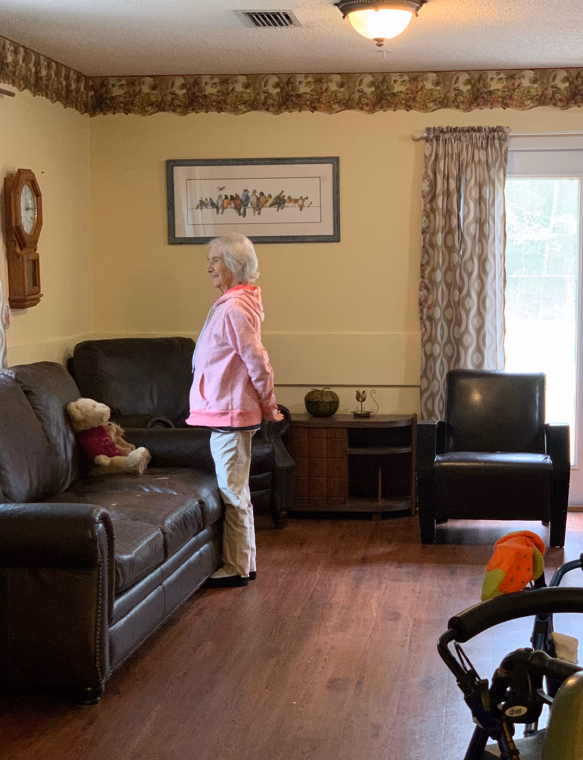A person stands beside a leather sofa in a living room with armchairs, framed bird artwork, a wall clock, and a walker visible.
