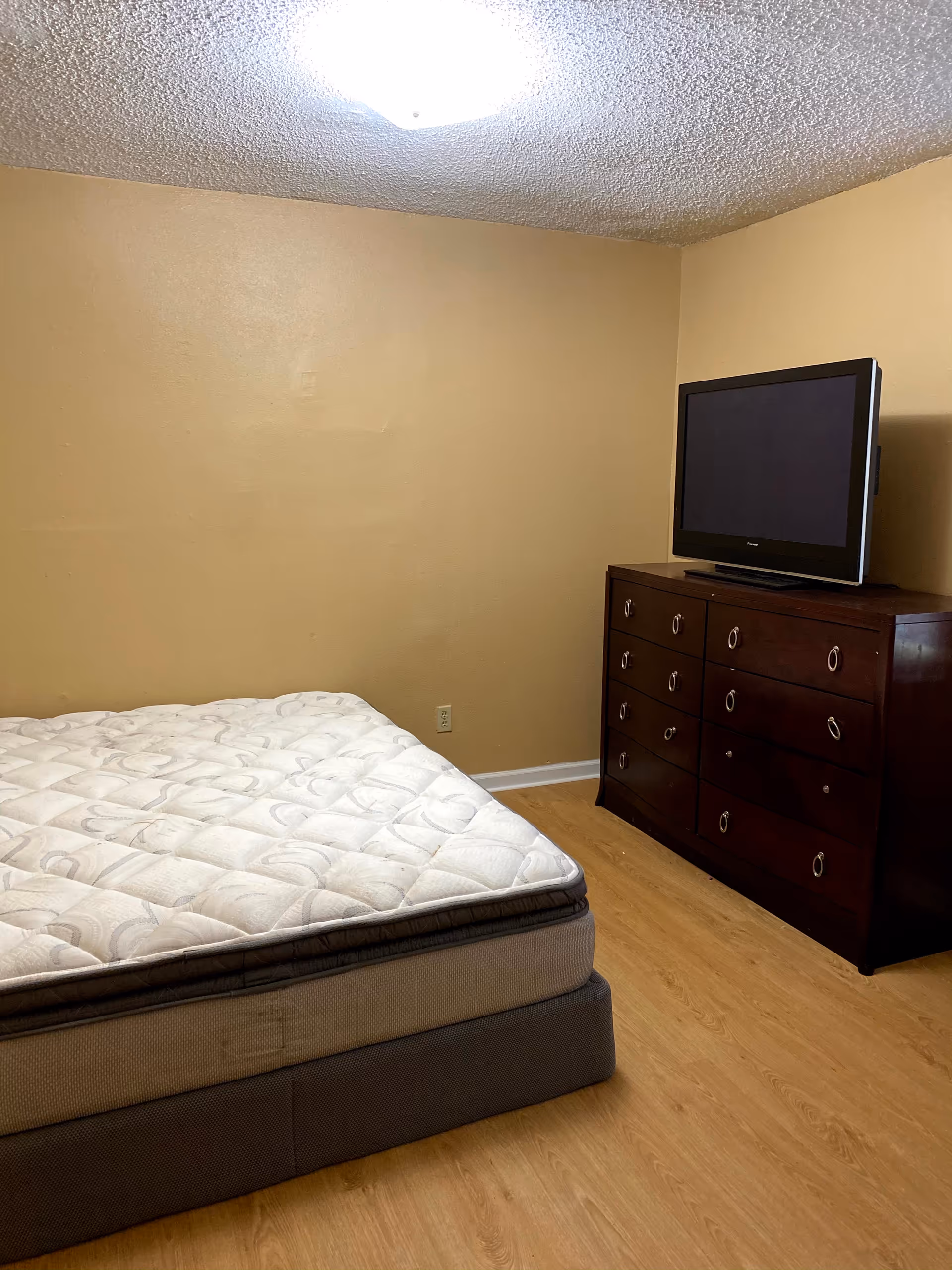 Unadorned bedroom with mattress, dresser, and TV in beige room.