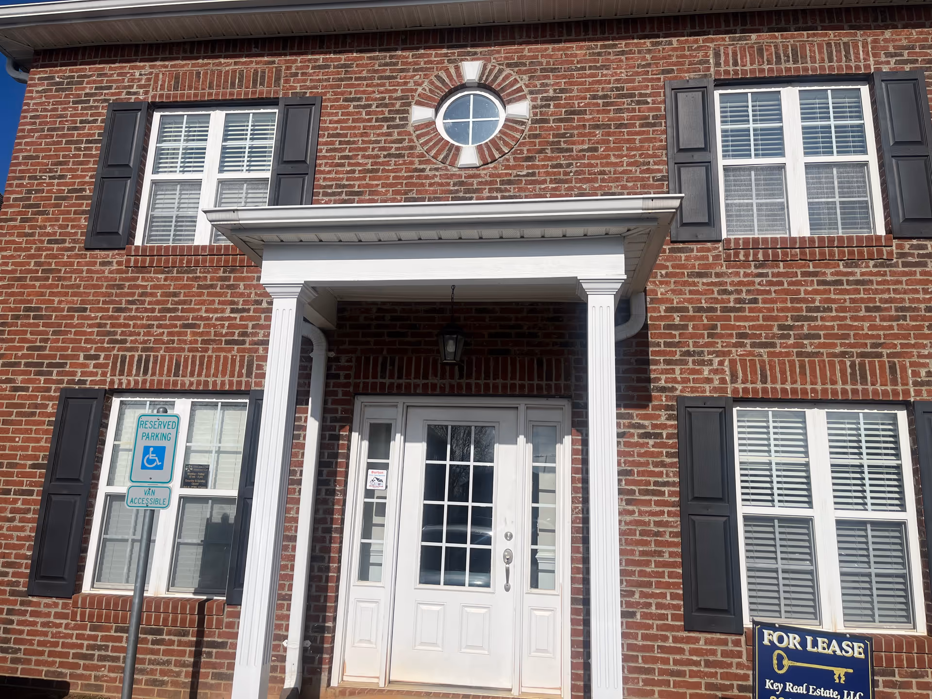 Brick building exterior with central white entrance, columns, round window above, and shuttered windows.