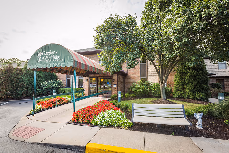 Entrance to Gardens at Westlake Senior Living facility with a green and red striped awning over the walkway, colorful flower beds on either side, a white bench, a small garden statue, and trees and shrubs surrounding the building.