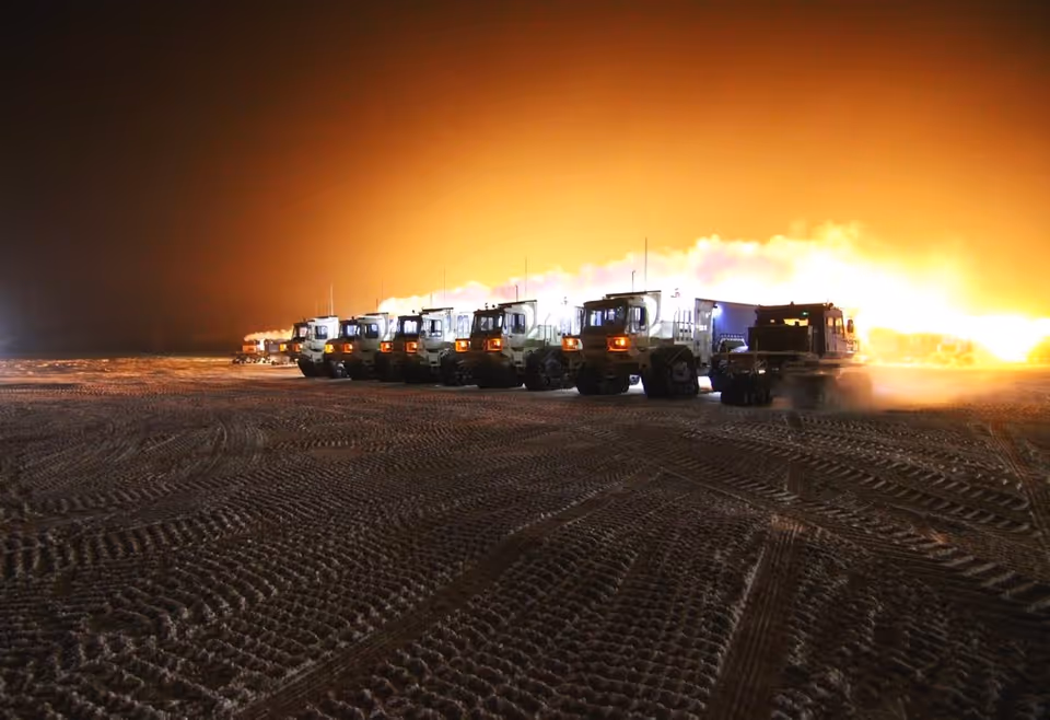A row of large trucks parked on a wide dark field with a huge fire behind.