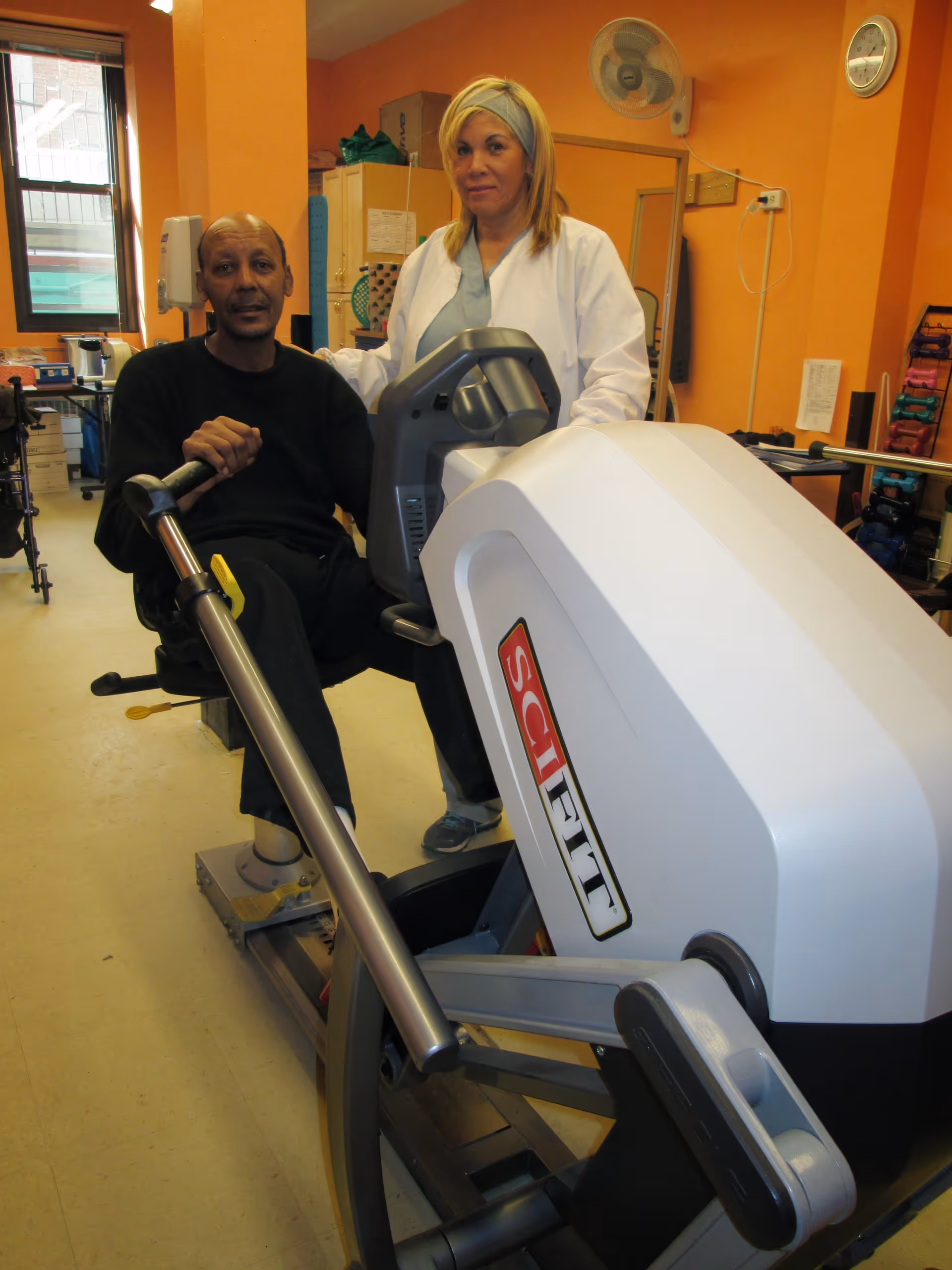A man using a SciFit exercise machine in a room with orange walls, accompanied by a woman in a white coat standing beside him. The room contains exercise equipment and has a window letting in natural light.