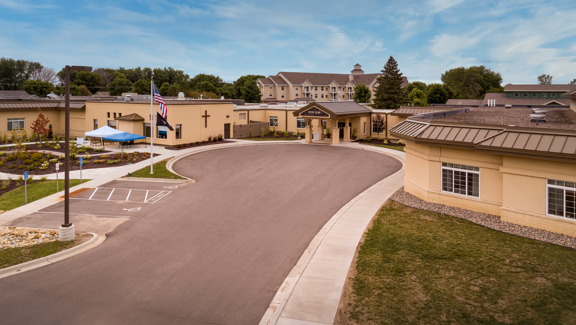An expansive senior living campus exterior with a curved driveway, beige buildings, and American flag.