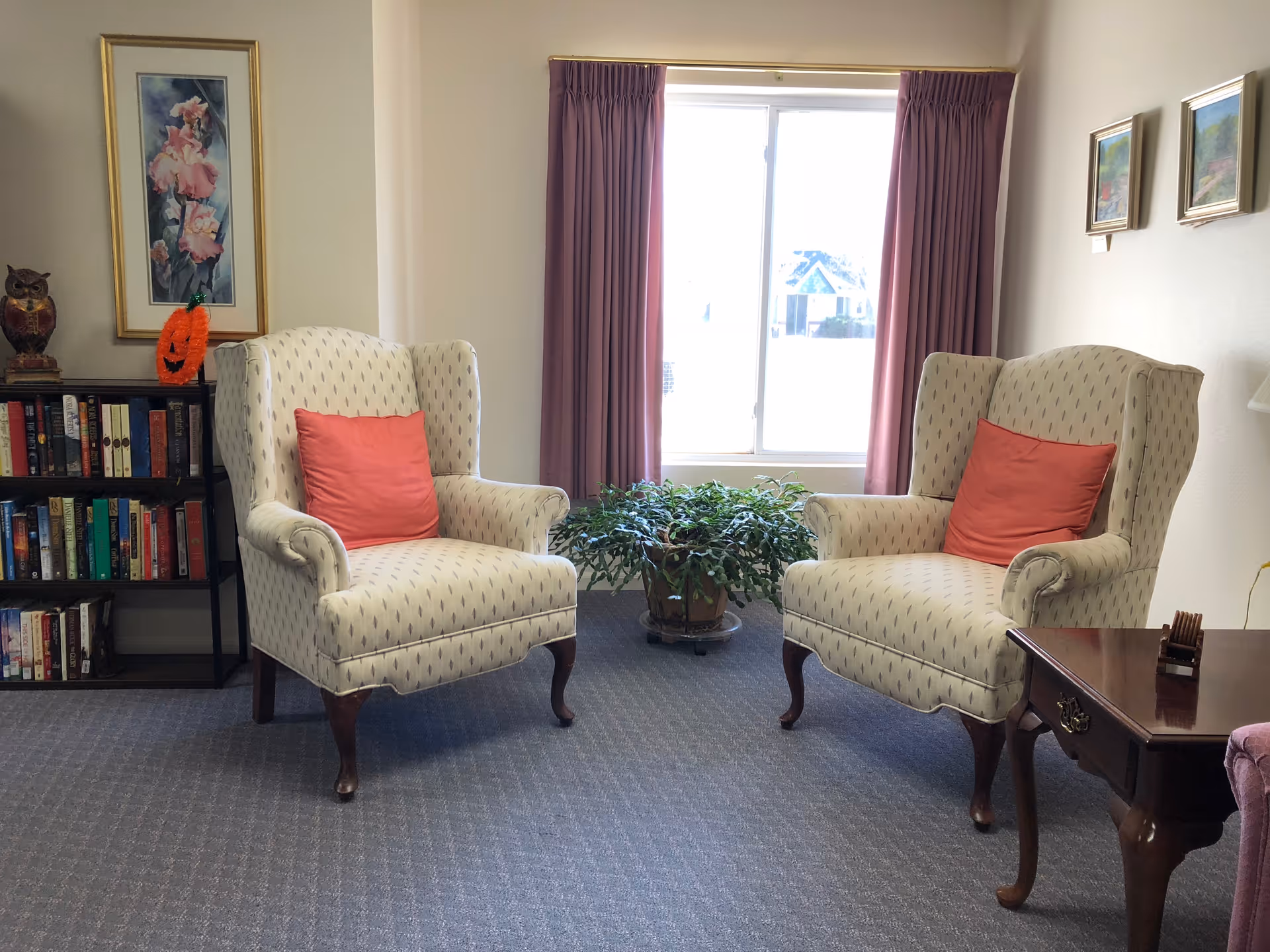A cozy sitting area with two upholstered armchairs featuring red cushions, positioned facing each other on a carpeted floor. Behind the chairs is a window with purple curtains and a green potted plant on a stand. To the left, there is a bookshelf filled with books and decorative items, including a framed floral painting and an owl figurine. On the right side, part of a wooden side table is visible with a small decorative item on it. The walls are light-colored with two small framed pictures hanging on the right.