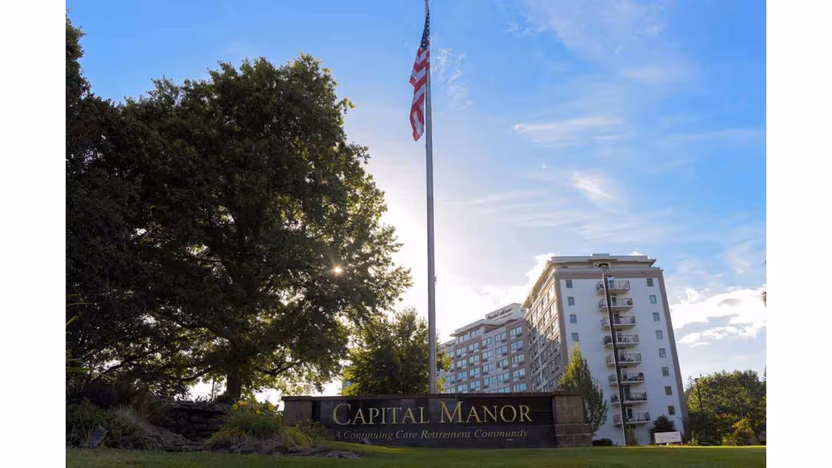 Exterior view of a multi-story retirement community building named Capital Manor with a large tree and an American flag on a flagpole in the foreground under a blue sky with some clouds.