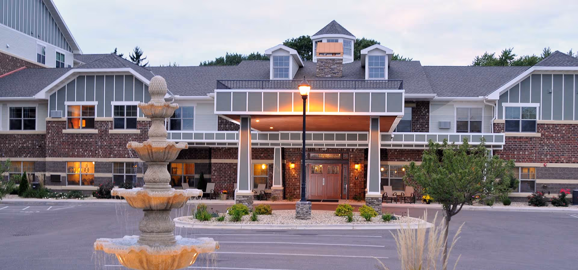 Front exterior view of Heritage Senior Campus building at dusk with a multi-tiered stone fountain in the foreground, a lit streetlamp, and landscaped greenery around the entrance.