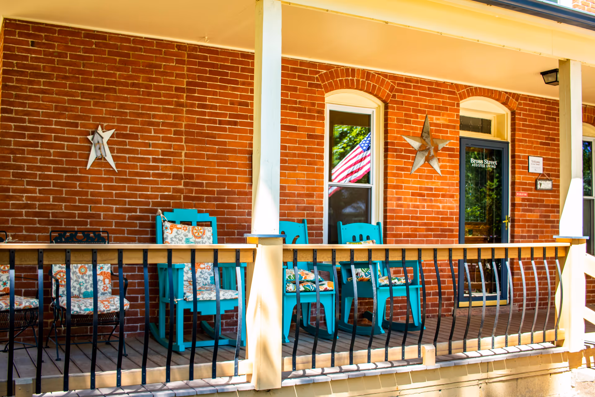Front porch of Bross Street Assisted Living with a brick wall, two decorative star ornaments, an American flag visible through a window, and several turquoise rocking chairs with floral cushions arranged along the porch railing.