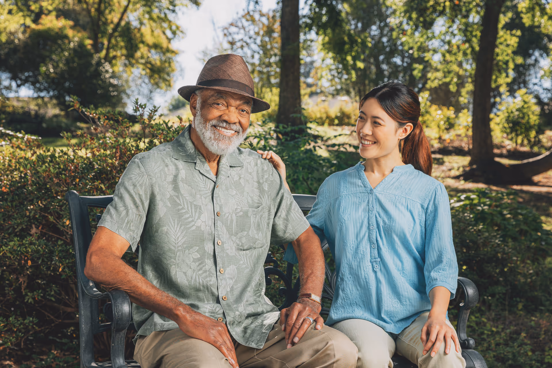 An elderly man and a female caregiver sit on a park bench outdoors, smiling.