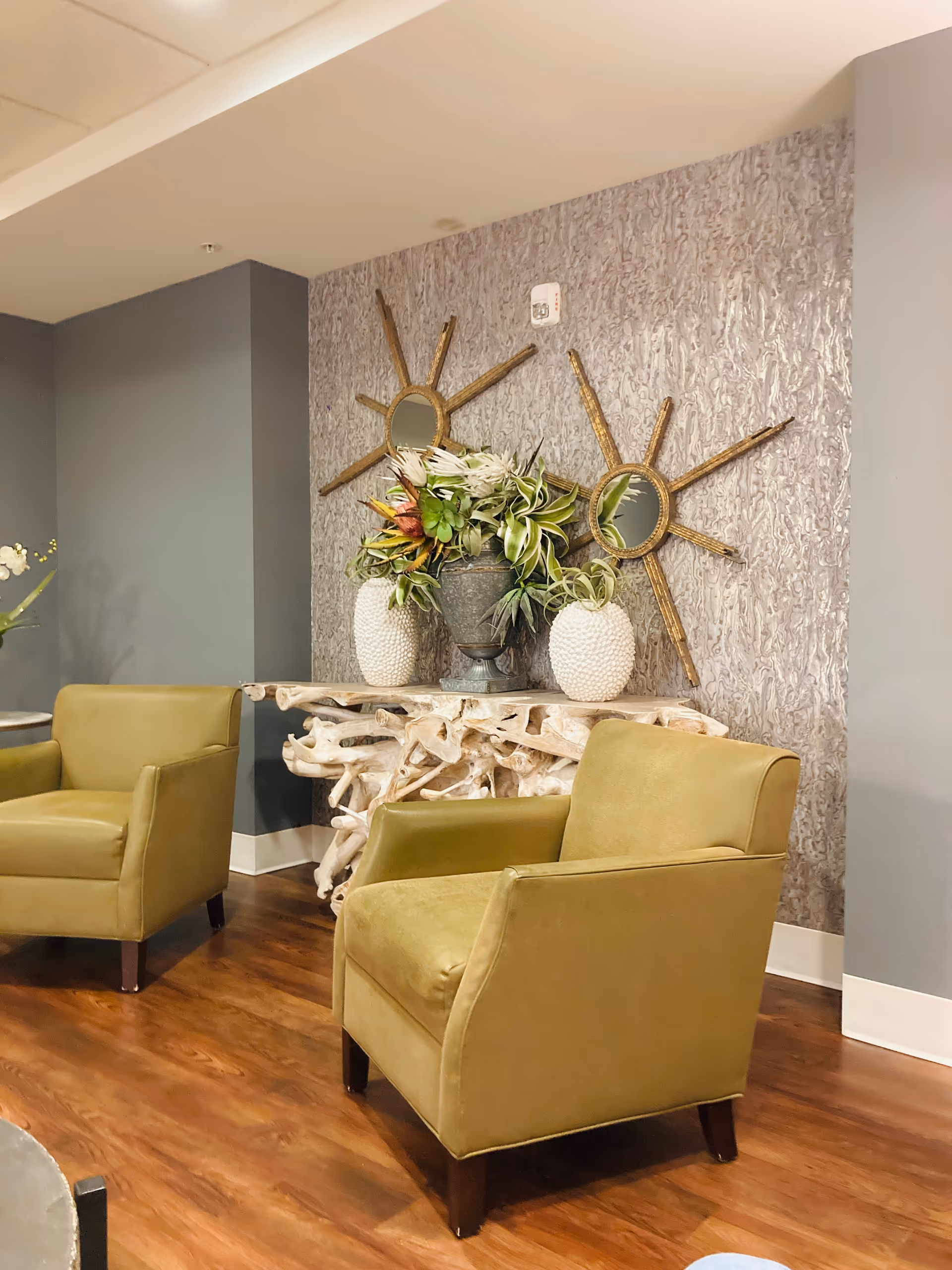 Soothing lounge area with olive chairs, a driftwood console table, plants, and sunburst mirrors.