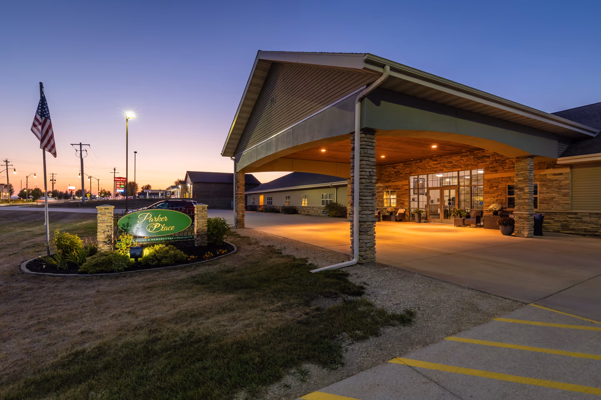 Exterior view of Parker Place Retirement Community at dusk, showing the entrance with a covered driveway supported by stone pillars, illuminated by warm lights. There is a landscaped area with a sign displaying the facility's name and an American flag on a pole nearby.