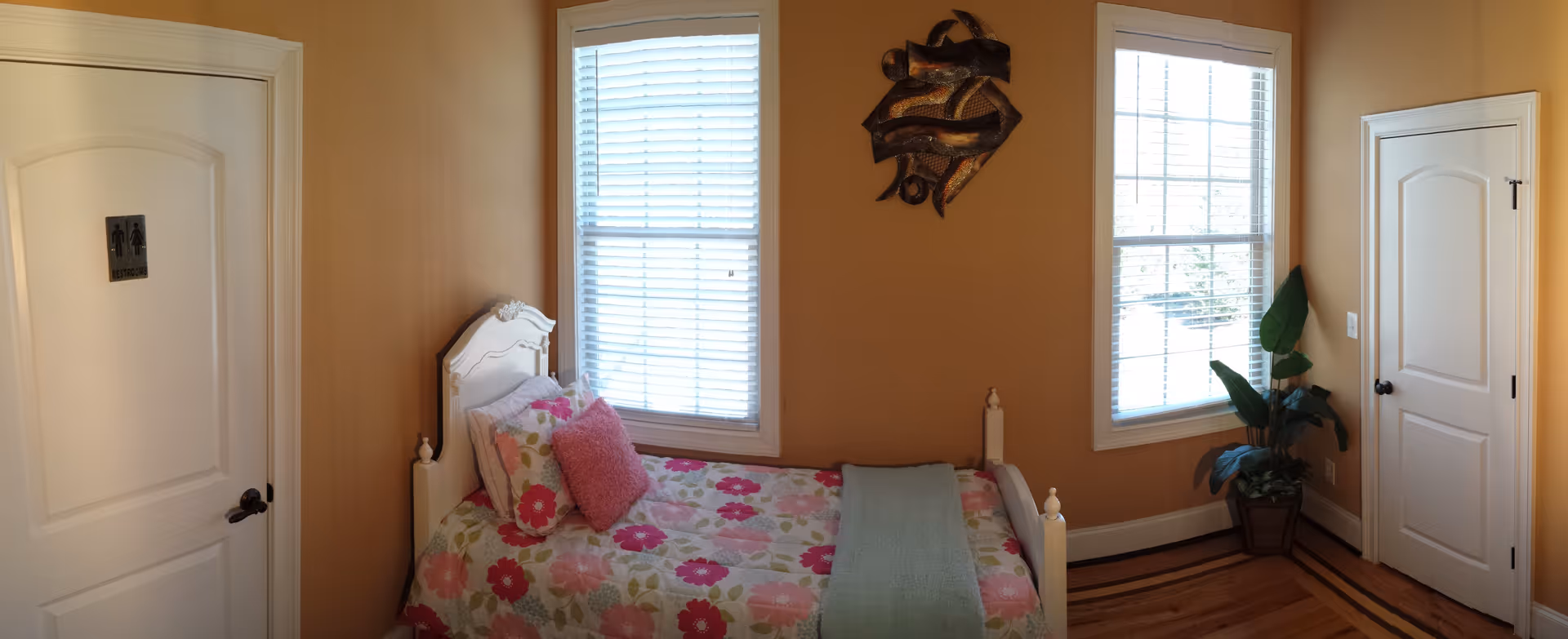 A small bedroom with beige walls featuring a single white bed with floral bedding and a pink pillow. There are two windows with white blinds, a decorative wall sculpture between the windows, a potted plant in the corner, and two white doors, one labeled as restrooms.
