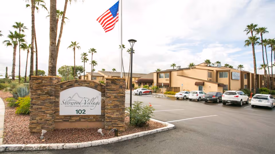 Exterior view of Sherwood Village assisted living building with the facility sign, American flag, palm trees, and a parking lot with cars.