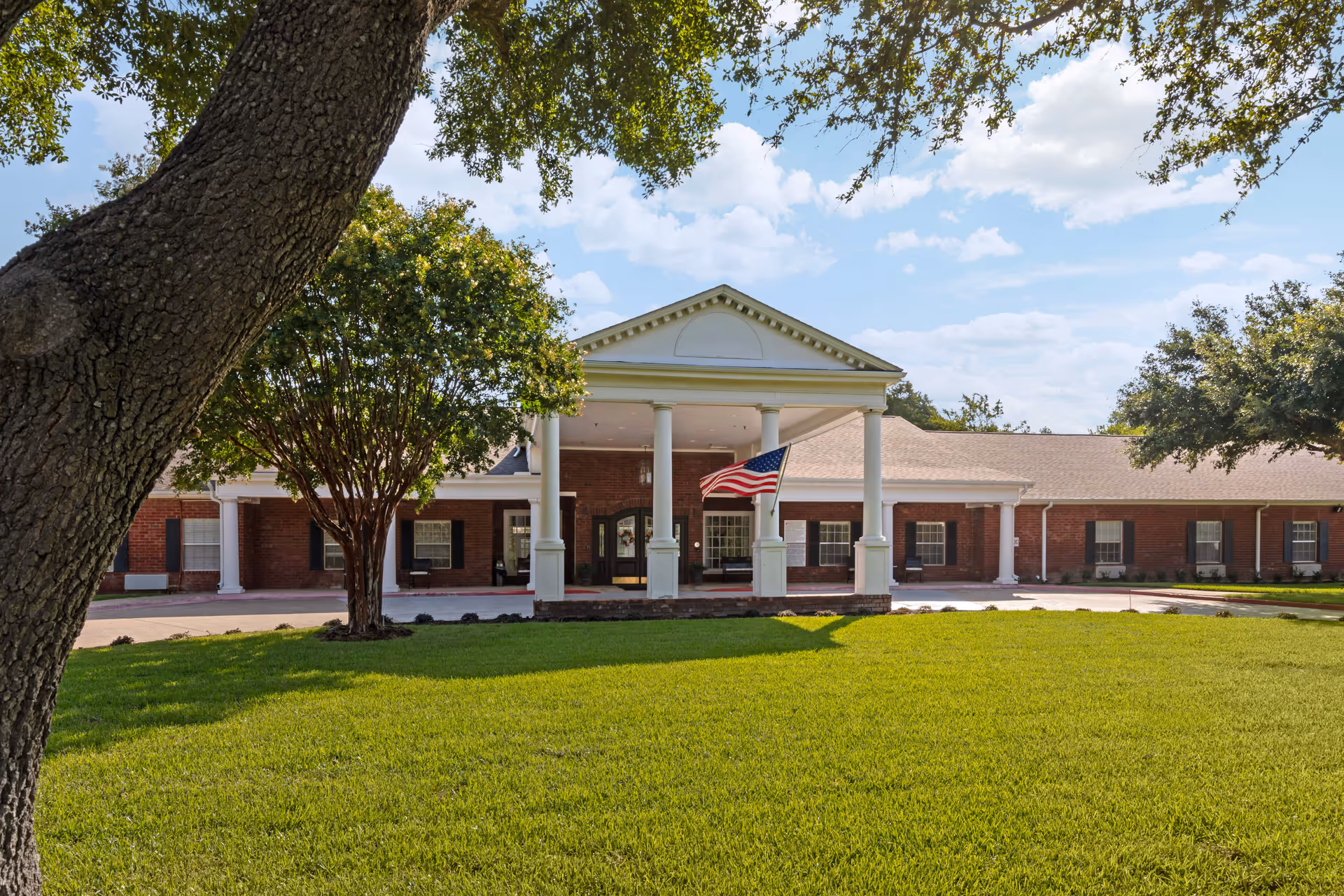 Front exterior view of Carver Heights Estates Senior Living building with a large green lawn, trees, and an American flag hanging from the white columned entrance under a blue sky with clouds.