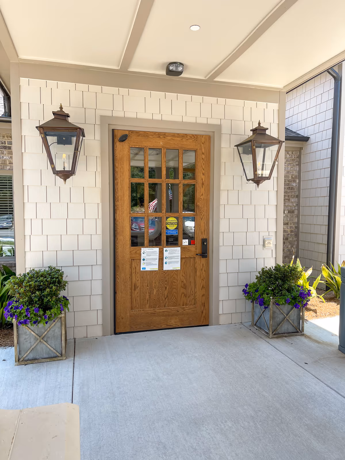 Wooden front door with glass panes, flanked by lanterns and planters on a light shingled exterior.