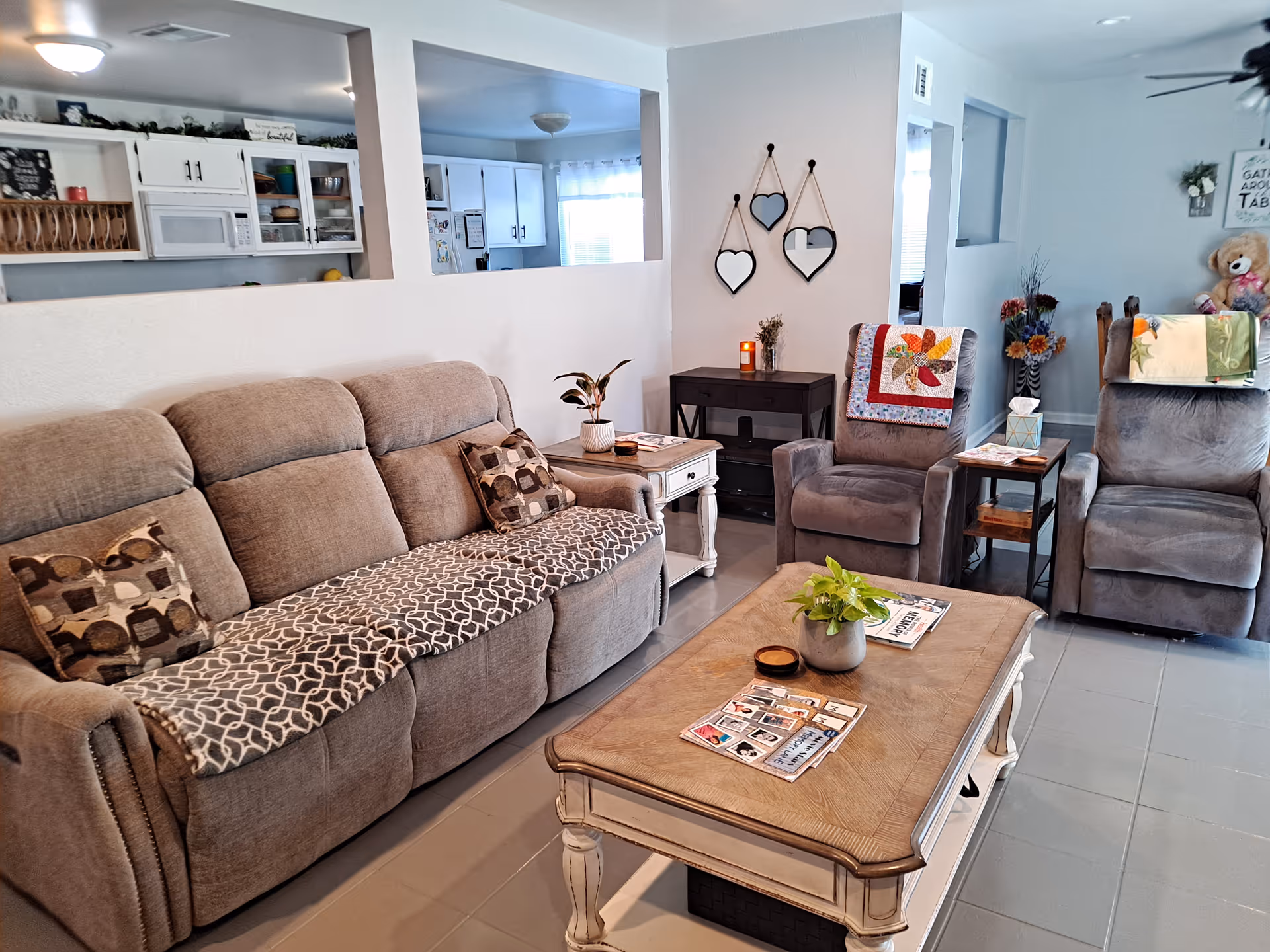 A cozy living room area with a beige sofa covered partially with a patterned throw, two gray armchairs with colorful blankets draped over the backs, a wooden coffee table with magazines and a small plant, and side tables with decorative items. The background shows a kitchen area with white cabinets and a window letting in natural light.