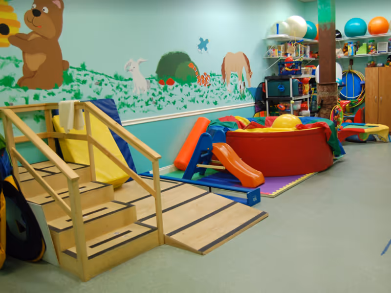 Colorful therapy playroom with a slide, soft mats, and wall mural of animals.