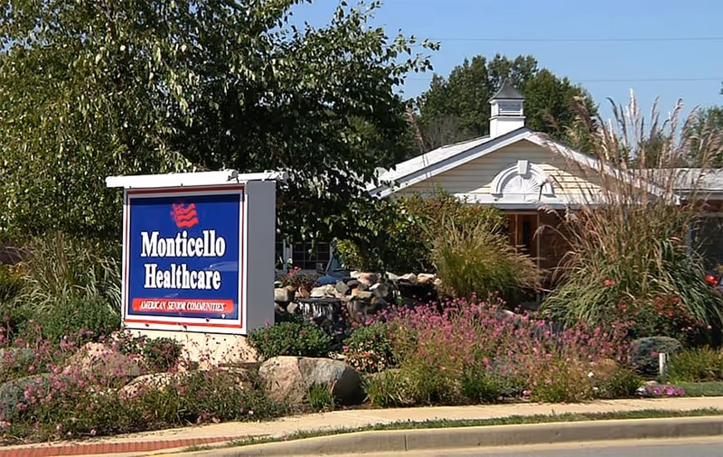 A Monticello Healthcare sign sits amid flowers and landscaping in front of the facility's single-story entrance.