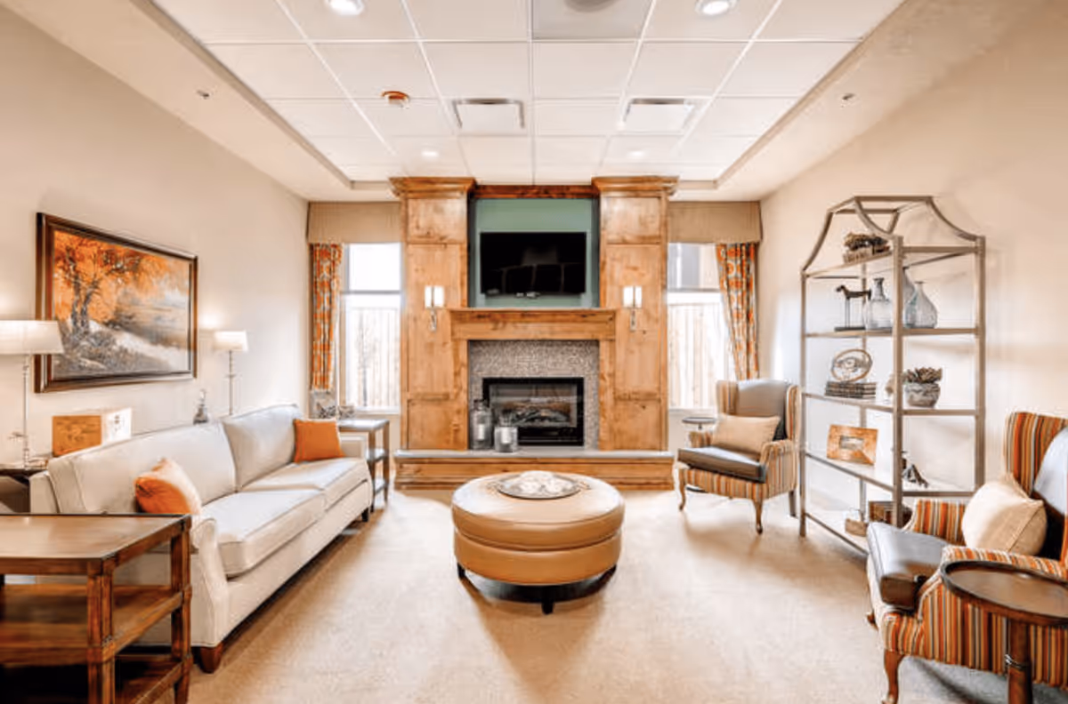 A cozy living room with a beige sofa adorned with orange and beige pillows, two striped armchairs, a round leather ottoman in the center, and a wooden fireplace with a mounted TV above it. The room has warm lighting, a large framed painting on the wall, two windows with patterned curtains, and a metal shelving unit with decorative items.