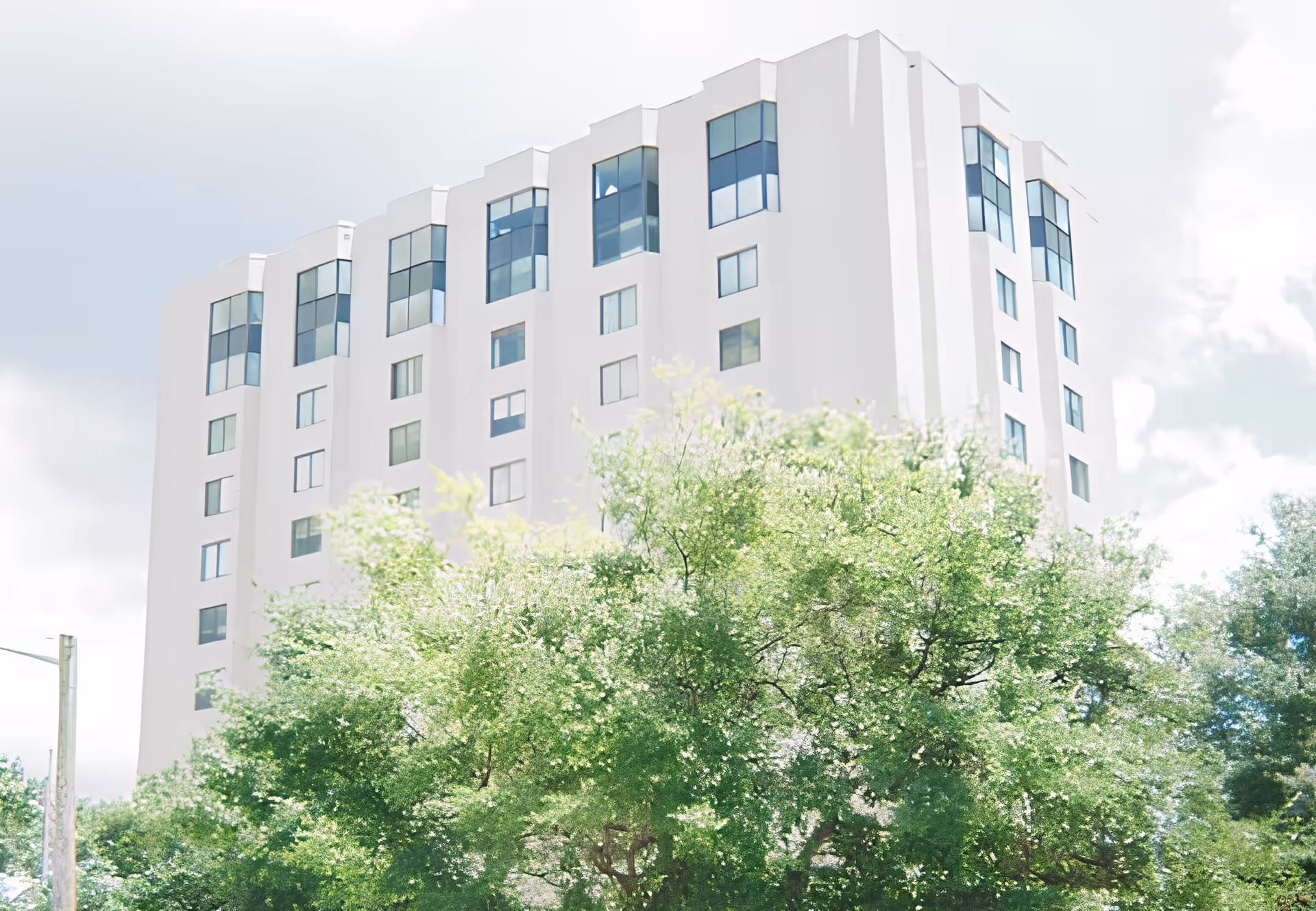 Tall white multi-story residential building rising above green trees under a cloudy sky.