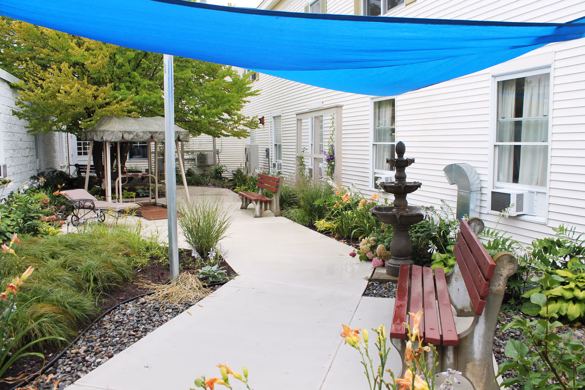 Outdoor garden area at a senior living facility with a concrete pathway, red benches, a multi-tiered fountain, various plants and flowers, a blue shade canopy overhead, and a gazebo swing in the background next to a white building.