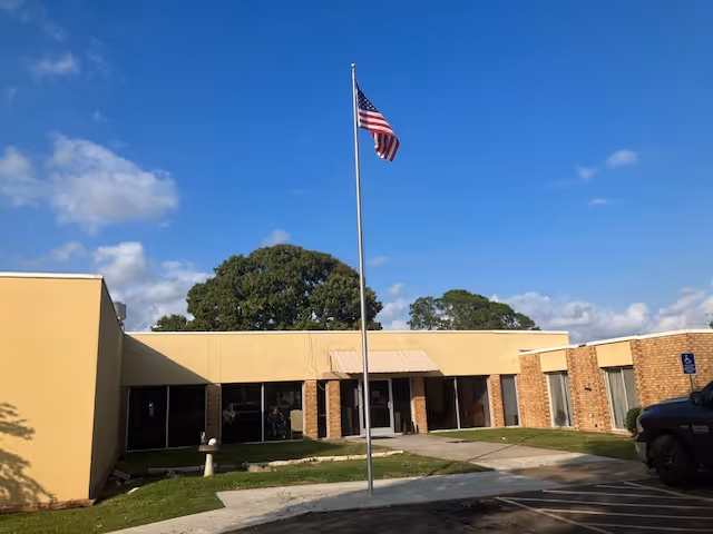 Single-story senior living building facade with an American flag on a pole in front under a blue sky.