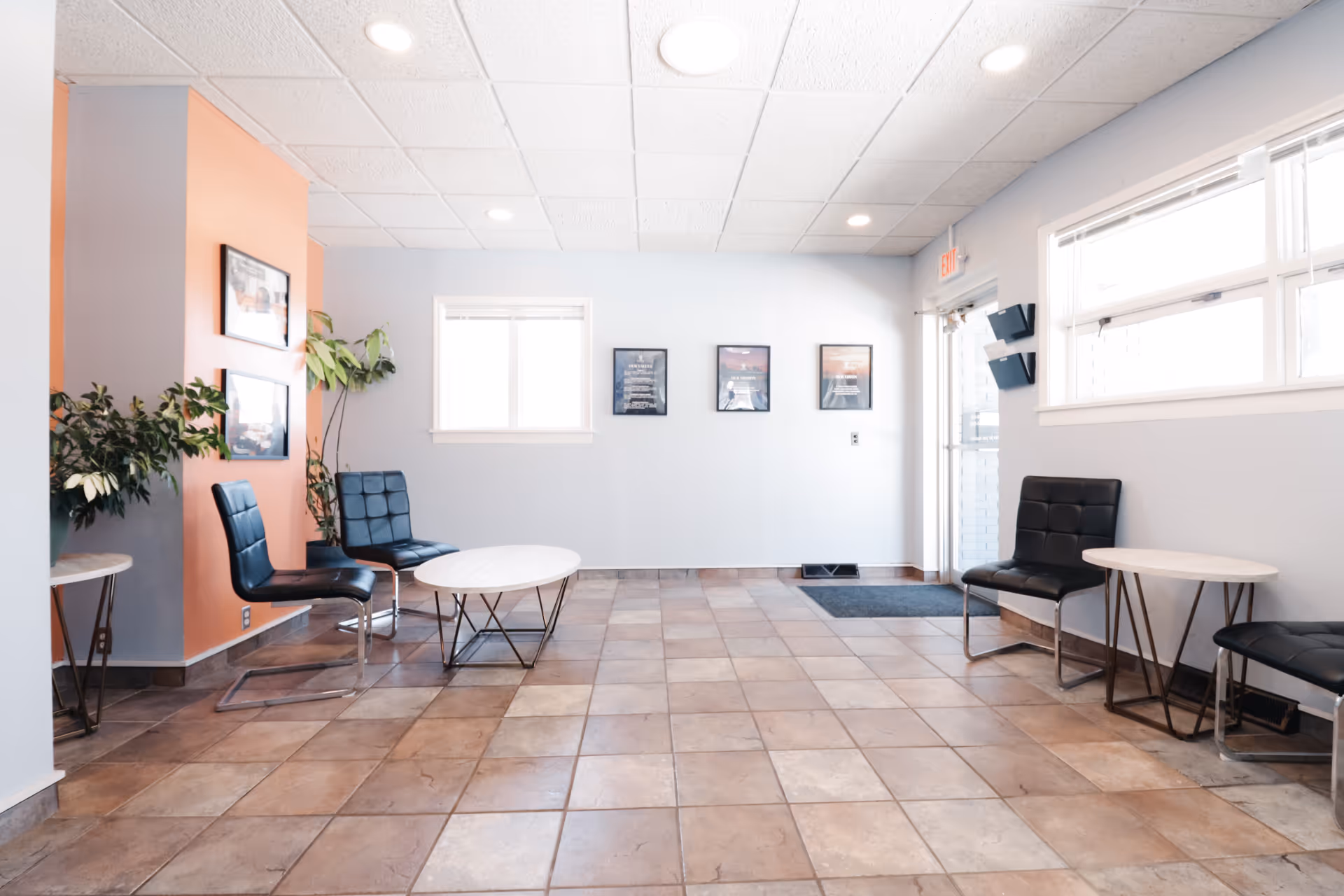 Interior waiting area with blue chairs, round tables, plants, and tiled flooring.